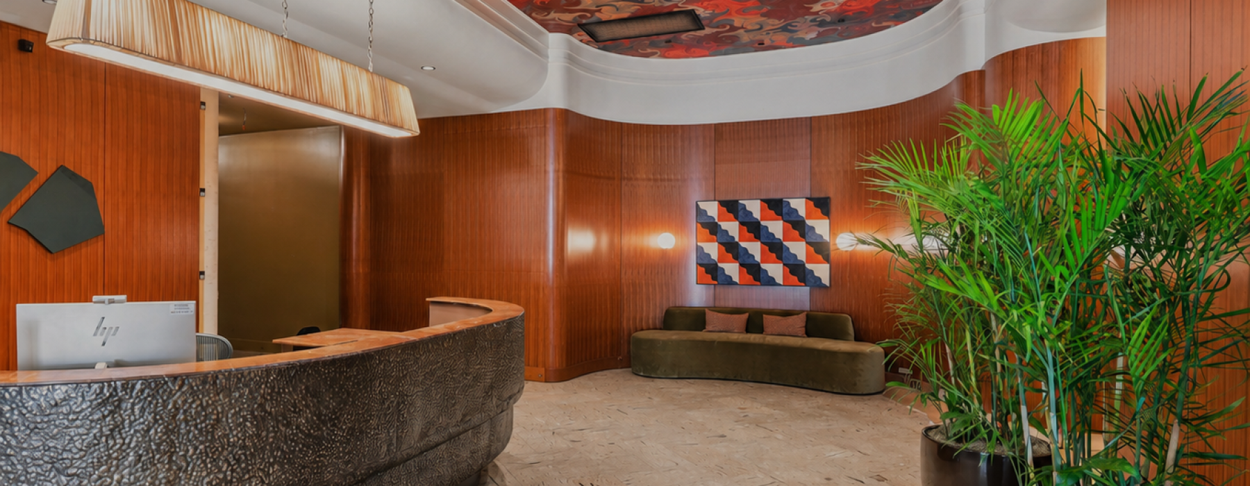 Modern apartment lobby at Huxley featuring a curved front desk, colorful patterned ceiling accent, lush indoor greenery, and seating.